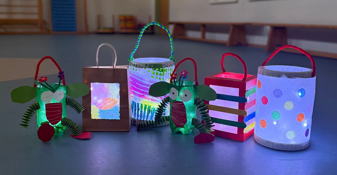 Vier bunte Bastelarbeiten auf einem Tisch: ein brauner Papierbeutel mit einer farbenfrohen Zeichnung, eine grüne Papierlaterne, eine rote Kastenlaterne und eine grüne Kreatur mit einem Griff.