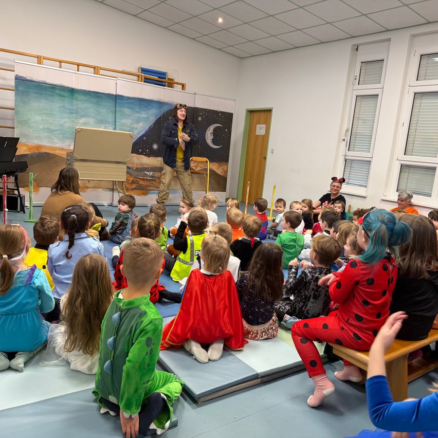 Ein Mann in einem Kostüm unterhält eine Gruppe von Kindern in verschiedenen Kostümen, die auf einem Teppich in einem Raum mit einer Wandmalerei und einer Holztür sitzen.