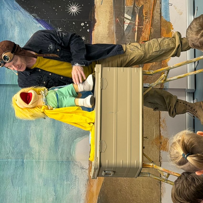 Ein Mann mit einer Pilotenbrille und einem Hut hält eine Puppe vor einer Metallbox, während Kinder zusehen.