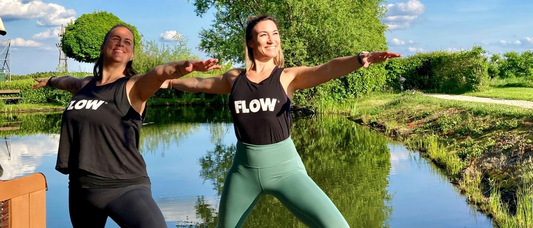 Zwei Frauen in schwarzen Tanktops mit 'FLOW' darauf machen Yoga auf einem Holzsteg am Teich.
