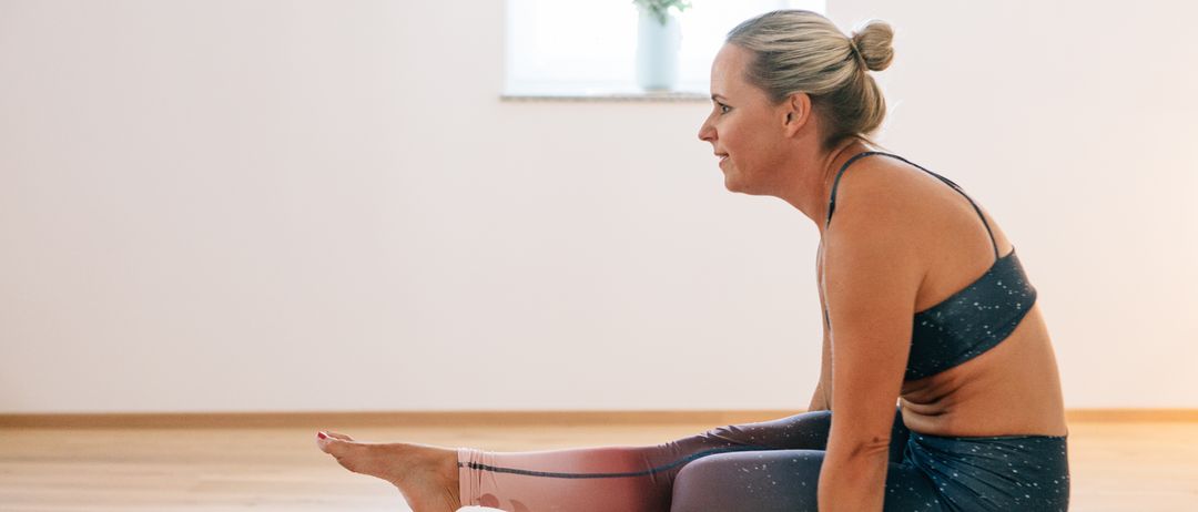 Eine Frau in Trainingskleidung kniet auf einem Holzboden, möglicherweise in einer Yoga-Pose, mit ihrem Fuß auf dem Boden.