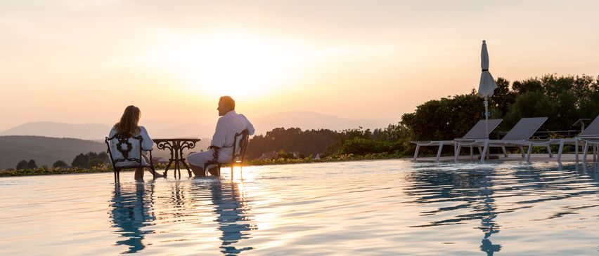 Ein Paar sitzt während des Sonnenuntergangs am Pool, einer in einem weißen Bademantel, der andere in einem Kleid. Ein kleiner Tisch trennt sie. Hinter ihnen stehen zwei zusammengeklappte Liegestühle, und ein geschlossener Regenschirm steht neben ihnen.