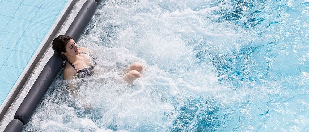Eine Frau entspannt sich in einem Schwimmbad, umgeben von Wasserspritzern, in einem schwarzen Badeanzug.