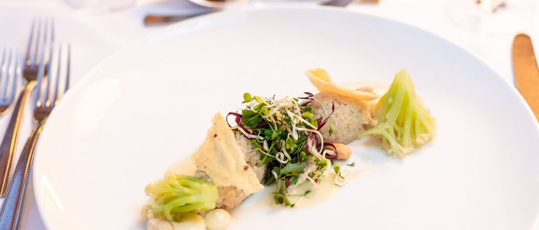 Ein Gourmetgericht mit einer Mischung aus grünem Gemüse, Bohnen und einem Reiskuchen. Der Teller steht auf einer weißen Oberfläche, mit Besteck und einem Glas im Hintergrund.
