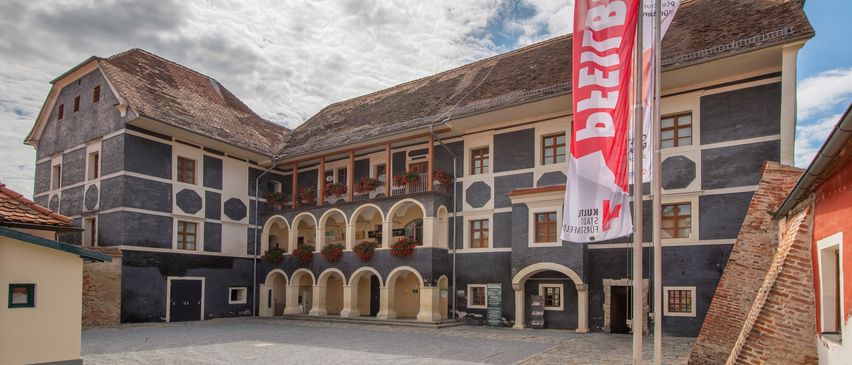 Ein historisches Gebäude mit einem Innenhof, gekennzeichnet durch eine große rote Fahne mit weißem Text, umgeben von Bogenfenstern und einem gepflasterten Innenhof unter einem bewölkten Himmel.