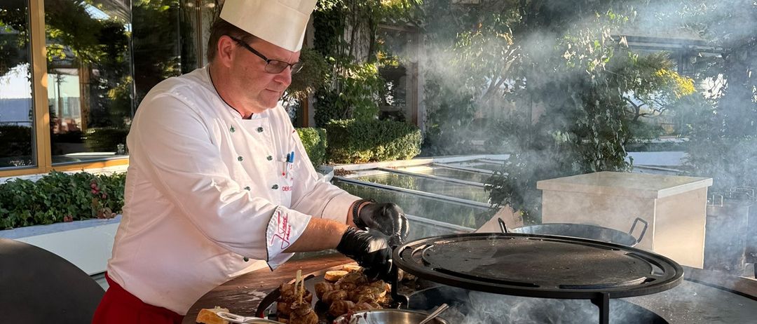 Ein Koch bereitet Fleisch auf einem Grill im Freien zu, trägt eine Kochmütze, Handschuhe und eine weiße Schürze. Rauch steigt vom Grill auf, und eine Schüssel und Utensilien sind in der Nähe.