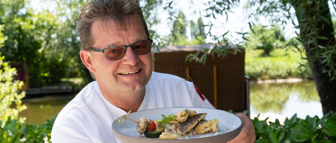 Ein Koch in einem weißen Hemd hält einen Teller mit einem Fischgericht, Gemüse und Garnierungen und lächelt.