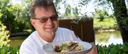 Ein Koch in einem weißen Hemd hält einen Teller mit einem Fischgericht, Gemüse und Garnierungen und lächelt.