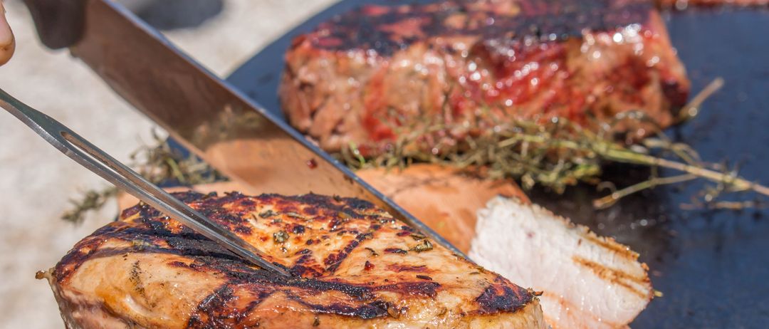 Eine Nahaufnahme eines Schweinekoteletts, das mit einem Messer auf einem Holzschneidebrett geschnitten wird. Kräuter und Rosmarin sind verstreut.