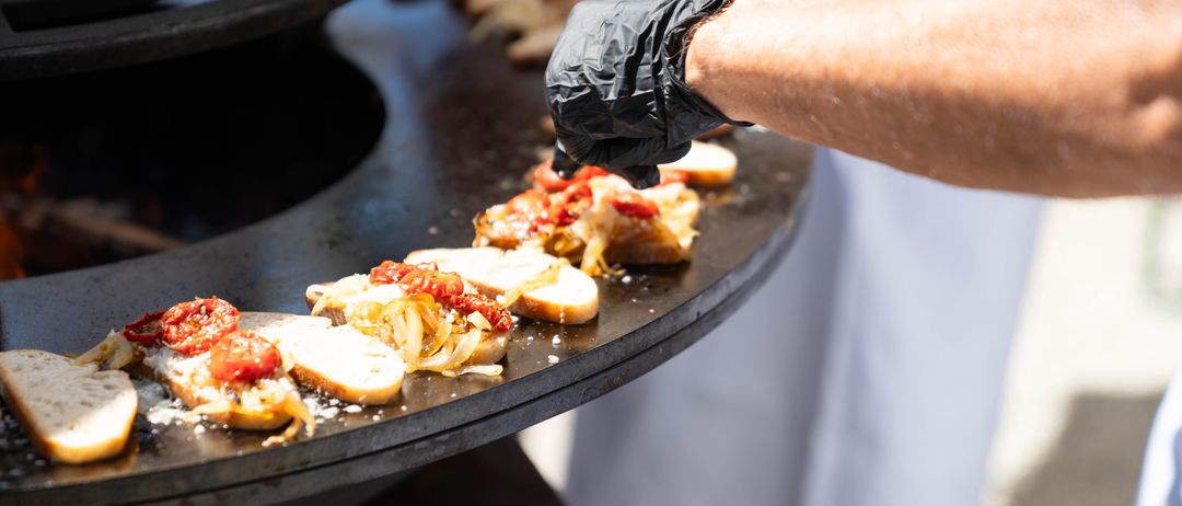 Mehrere Personen bereiten Essen auf einem Grill zu. Sie tragen weiße Schürzen und schwarze Handschuhe. Das Essen besteht aus Brot mit Toppings wie Tomaten und Zwiebeln.