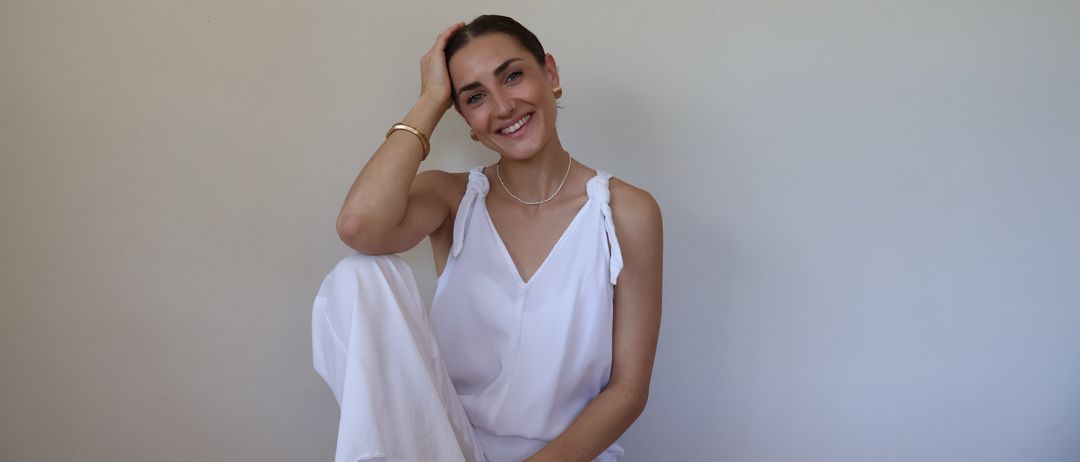 Eine Frau in einem weißen Kleid und Accessoires sitzt gegen eine weiße Wand, lächelt und hält ihren Kopf.