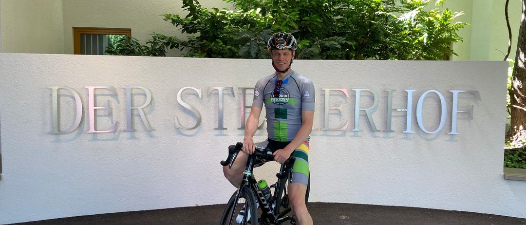 Ein Radfahrer mit Helm posiert für ein Foto vor einer weißen Wand mit dem Namen 'Der Steinberghof'.