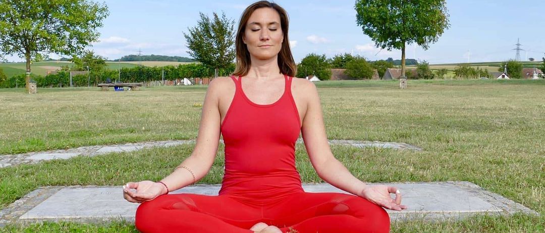 Eine Frau in rotem Outfit sitzt auf dem Rasen in einer Yoga-Pose. Sie befindet sich in einer ruhigen Außenumgebung mit Bäumen und einem klaren blauen Himmel.
