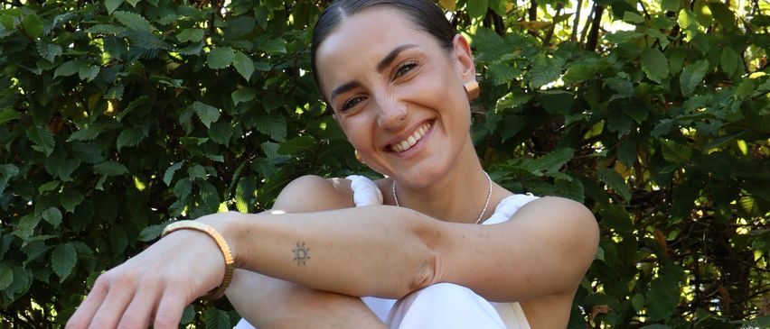A smiling woman poses outdoors in front of greenery, wearing a white dress and gold bracelets, with a tattoo on her left arm.