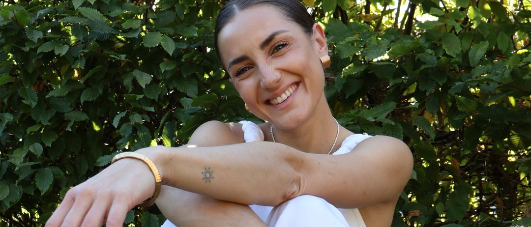 Eine lächelnde Frau posiert im Freien vor Grünpflanzen, trägt ein weißes Kleid und goldene Armbänder, mit einer Tätowierung am linken Arm.