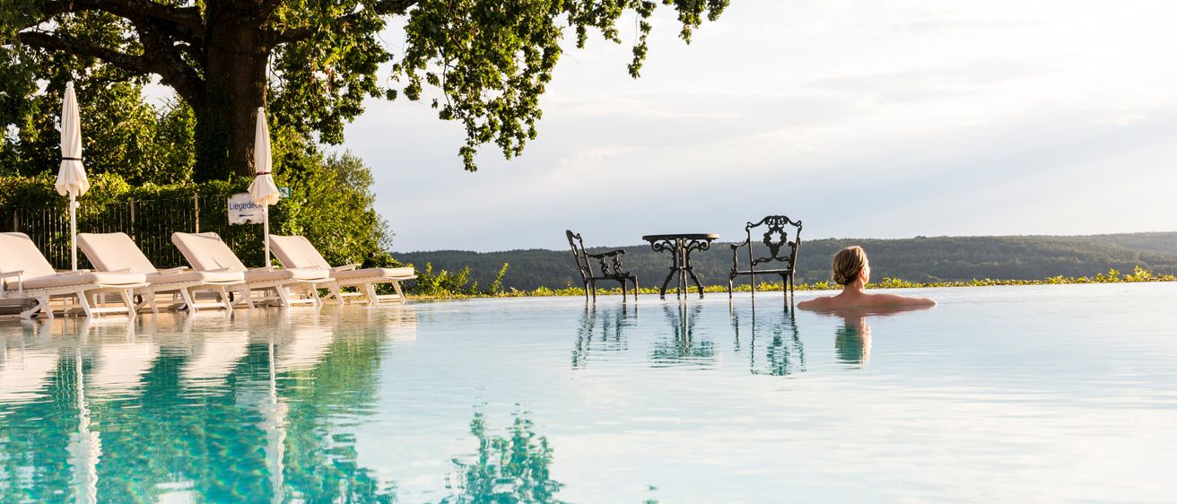 Eine ruhige Poolszene mit einer schwimmenden Frau, umgeben von Stühlen, einem Tisch und Liegestühlen. Bäume und Berge sind im Hintergrund.
