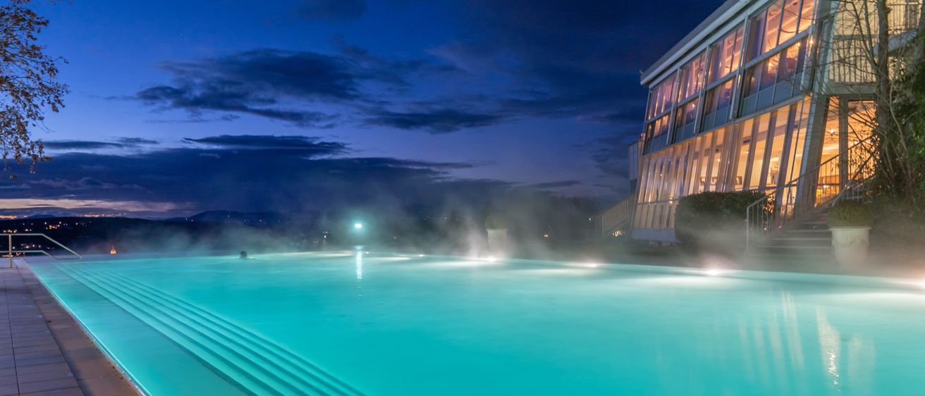 Ein großer Swimmingpool bei Nacht mit einem Gebäude und einem hellen Licht. Das Wasser ist blau und reflektiert den Himmel.