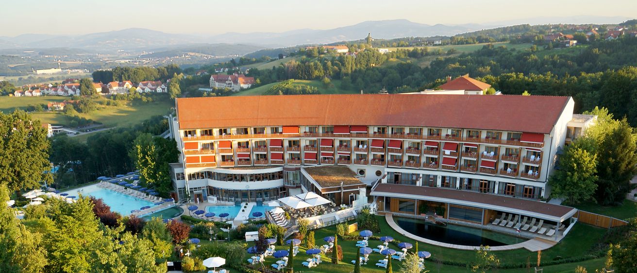 Ein Luftbild eines Resorthotels mit mehreren Schwimmbädern, einem Springbrunnen und Sonnenschirmen. Das Gebäude hat mehrere Stockwerke und rote Dächer.