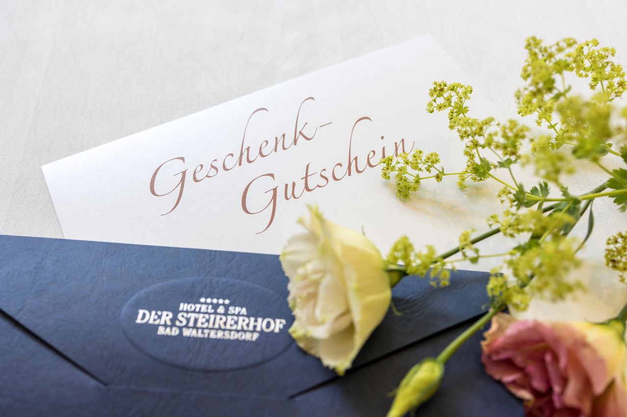 Ein Geschenkgutschein vom Hotel und Spa Der SteirerhoF in Bad Waltersdorf liegt auf einem blauen Umschlag. Das weiße Papier hat geschwungenen Text, der 'Geschenk-Gutschein' lautet. Eine gelbe Rose liegt auf dem Umschlag.