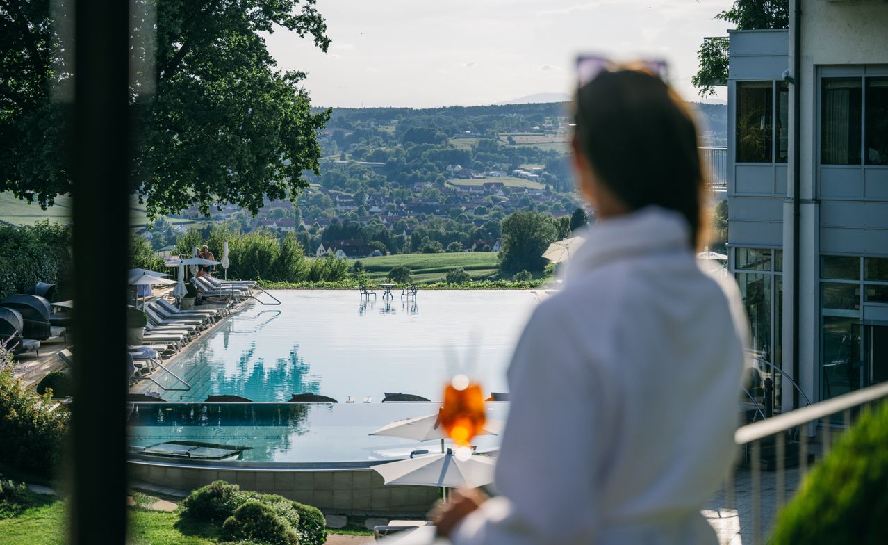 Eine Frau in einem weißen Bademantel steht am Rande eines großen Pools und blickt auf eine friedliche Landschaft mit einer Stadt in der Ferne.