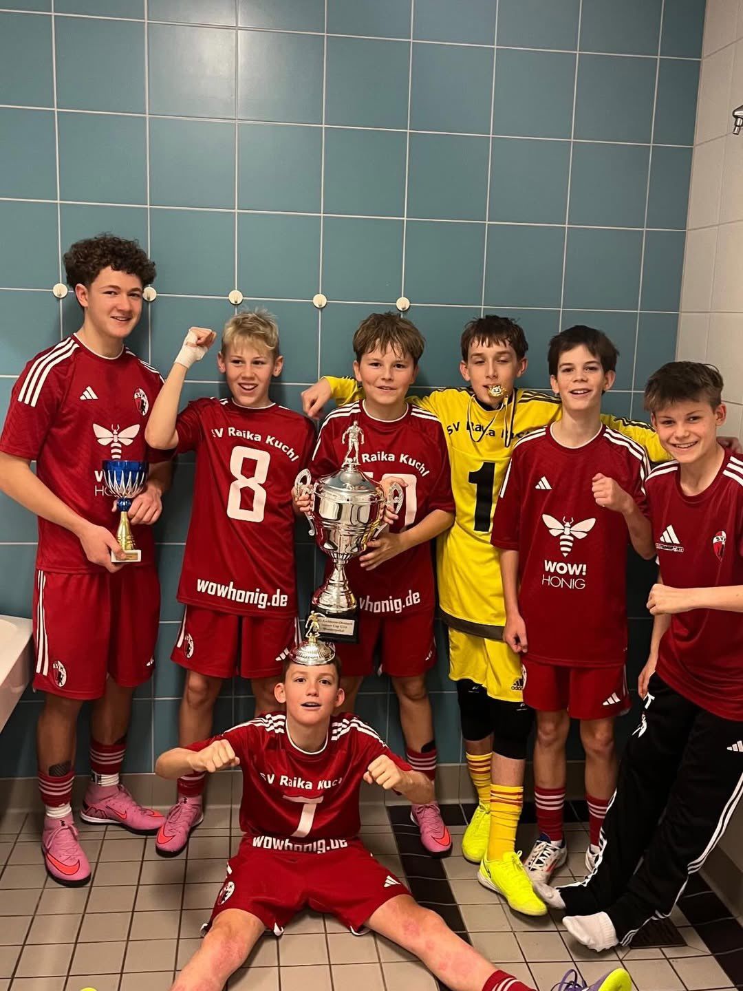 Eine Gruppe junger Fußballspieler, alle in roten Trikots, posiert mit ihren Trophäen. Ein Junge hält eine Goldmedaille. Das Trikot des Teams zeigt ein Bienenlogo.