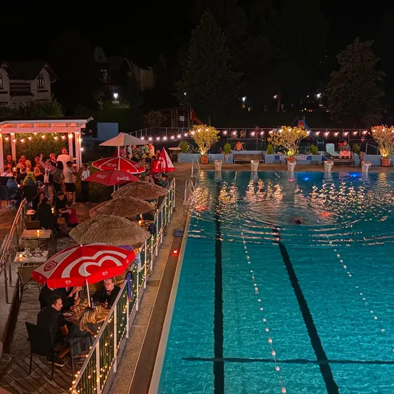 Eine nächtliche Szene an einem Poolbereich mit Menschen, die unter Schirmen essen, einem Schwimmbecken mit blauen Lichtern und einer festlichen Atmosphäre mit Lichterketten.