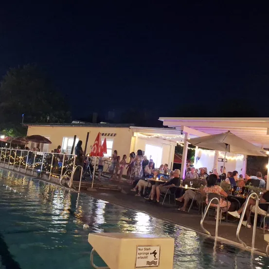 Eine Nachtszene in einem Außenpoolbereich mit Menschen, die um Tische unter Sonnenschirmen sitzen, einige stehen am Pool, und ein Gebäude mit Lichtern und Fahnen im Hintergrund.