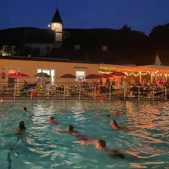 Eine Gruppe von Menschen schwimmt nachts in einem Pool, mit einem Gebäude und einem Turm im Hintergrund.