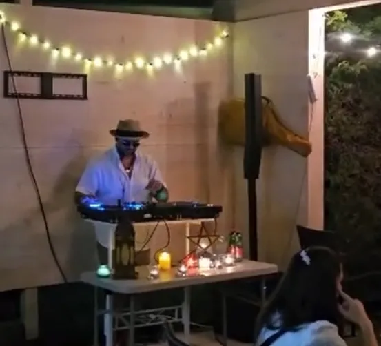 Ein Mann mit Hut und Sonnenbrille spielt Musik auf einer DJ-Anlage in einem schwach beleuchteten Raum mit Lichterketten und einer Frau, die in der Nähe sitzt.