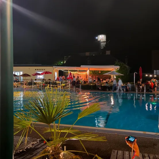 Ein nächtlicher Blick auf einen Poolbereich, wo Menschen an Tischen unter Sonnenschirmen essen, beleuchtet von Lichterketten und einer hellen Lampe.