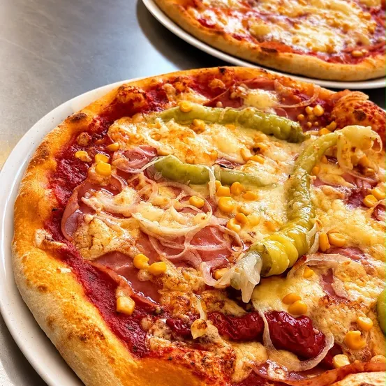 Ein Nahaufnahme einer Pizza mit Toppings wie Mais, grünen Paprika, Zwiebeln und Schinken auf einem weißen Teller. Eine weitere Pizza ist im Hintergrund teilweise zu sehen.