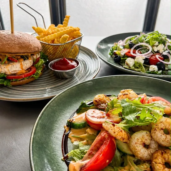 Drei Teller mit Essen auf einem Tisch, darunter ein Hähnchenburger, ein Salat mit Garnelen und ein Korb mit Pommes und Ketchup.