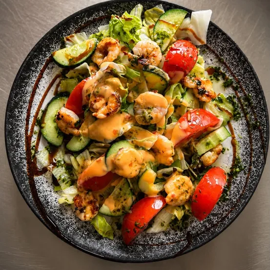 Ein Teller Salat mit gegrillten Garnelen, Tomaten, Gurken und einer Saucenüberzug auf grauem Hintergrund.