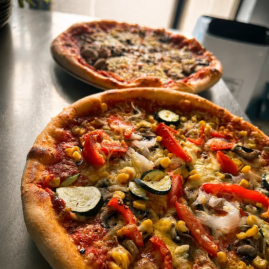 Zwei Pizzen mit verschiedenen Belägen wie Pilzen, Mais und Paprika auf einer Metalloberfläche.