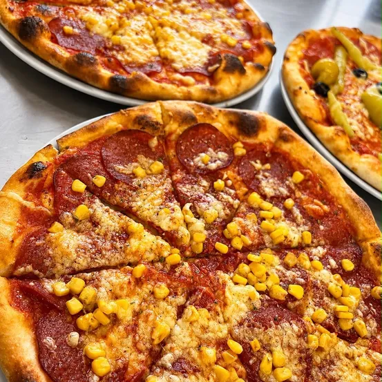 Drei Pizzen auf weißen Tellern mit Belägen wie Peperoni, Mais und Oliven. Die Pizza im Vordergrund hat Mais und Peperoni.