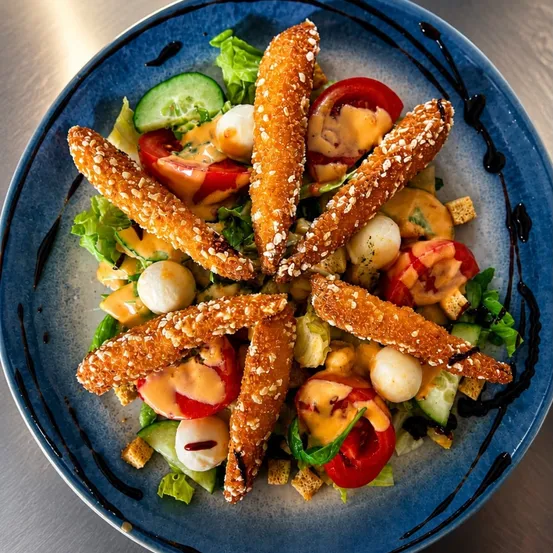 Ein blauer Teller mit einem Salat bestehend aus panierten Hähnchenstreifen, Salat, Tomaten, Gurken und einer cremigen Vinaigrette.