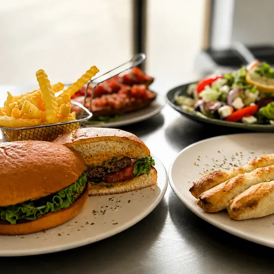 Ein Tisch mit verschiedenen Speisen, darunter Burger, Pommes, Salat und Ciabatta. Die Burger sind auf weißen Tellern, Pommes in einem Korb, Salat auf einem grünen Teller und Ciabatta auf einem weißen Teller.