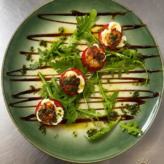 Ein grüner Teller mit vier mit Garnelen gefüllten Kirschtomaten, überzogen mit Käse und Kräutern, garniert mit Rucola und mit brauner Sauce übergossen.