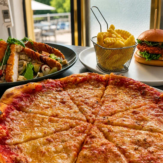 Ein Tisch mit einer großen Pizza, einem Salat und einer Frittenschale. Ein Burger ist ebenfalls auf dem Tisch.