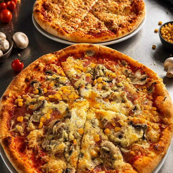 Zwei Pizzen auf einem Tisch mit Belägen wie Käse, Mais, Pilzen und Schinken. Eine Pizza hat ein fehlendes Stück.