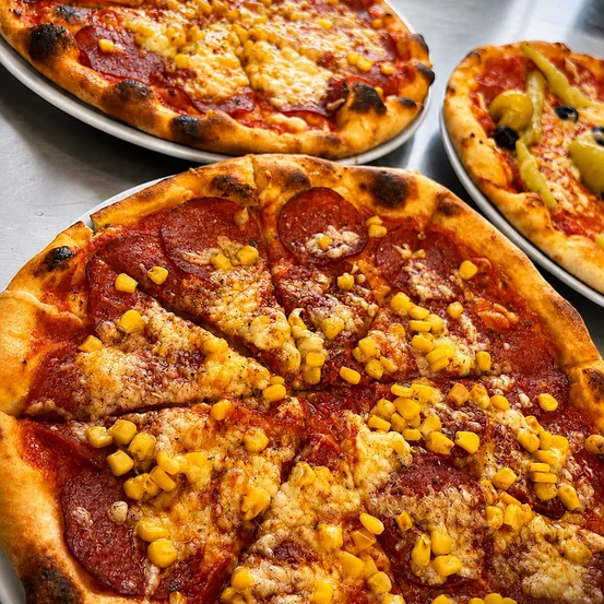 Drei Pizzen auf weißen Tellern mit Belägen wie Peperoni, Mais und Oliven. Die Pizza im Vordergrund hat Peperoni und Mais.