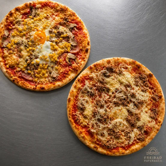 Zwei Pizzen auf einer metallischen Oberfläche. Eine hat ein gebratenes Ei, Mais und Pilze. Die andere hat Zwiebeln, Fleisch und Käse.