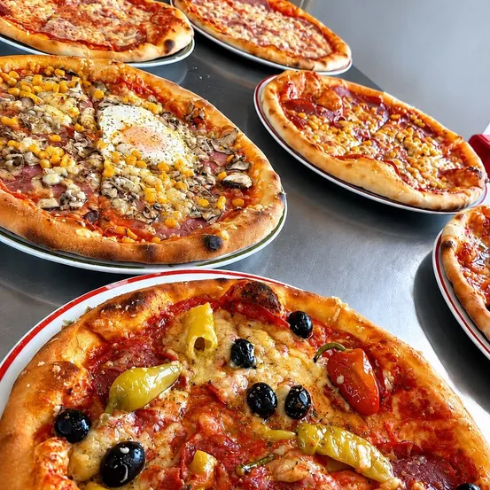Eine Auswahl an Pizzen ist auf einem Tisch ausgestellt, darunter eine Pizza mit gebratenem Ei, Mais und Pilzen und eine weitere mit Oliven, Tomaten und Paprika.
