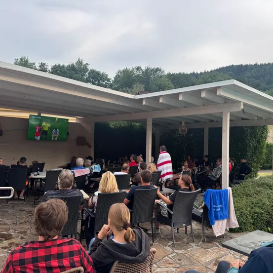 Eine Gruppe von Menschen sitzt unter einem Pergola und schaut sich ein Fußballspiel auf einem Fernseher an. Der Bereich ist von Grün und Bäumen umgeben.