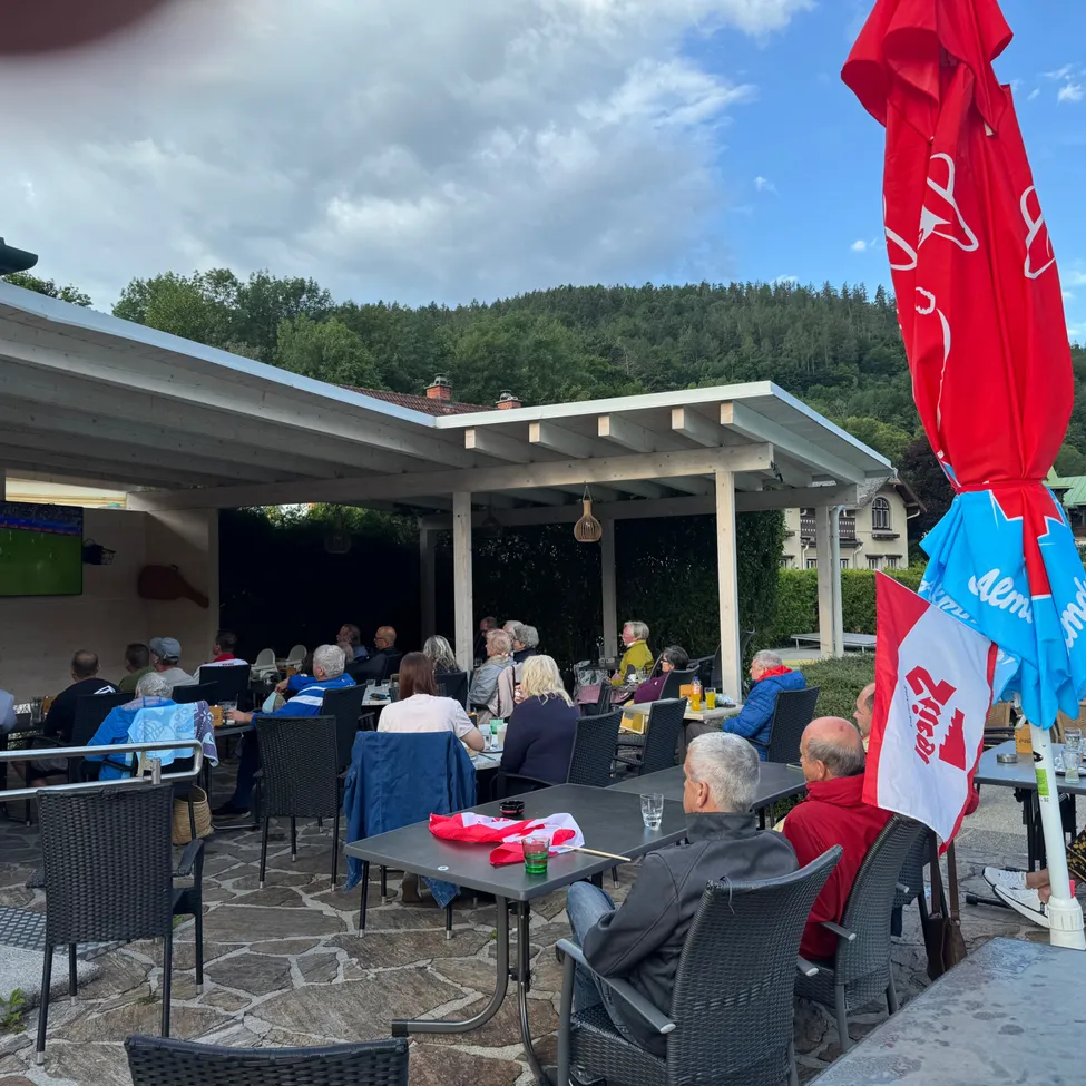 Eine Gruppe von Menschen sitzt unter einem überdachten Patio um Tische herum, mit Bergen und Bäumen im Hintergrund. Einige tragen Jacken und Mützen, und eine Person hält eine Flagge.