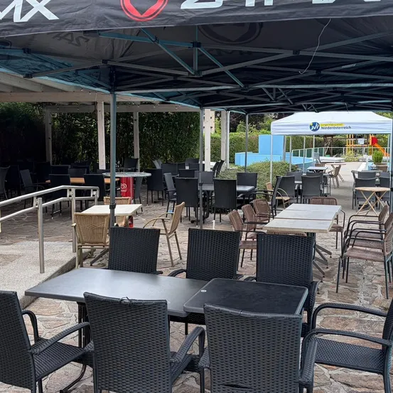 Ein Außenbereich eines Restaurants mit Tischen und Stühlen unter einem großen Zelt, das ein rotes Logo auf dem Baldachin aufweist. Der Bereich ist leer, mit einigen Tischen und Stühlen in Reihen angeordnet. Im Hintergrund befindet sich ein weißes Zelt und ein Poolbereich.