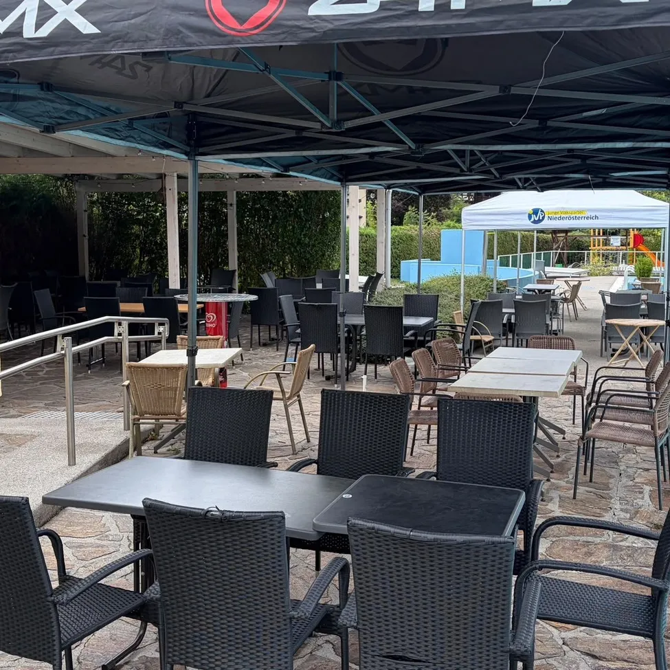 Ein Außenbereich eines Restaurants mit Tischen und Stühlen unter einem großen Zelt, das ein rotes Logo auf dem Baldachin aufweist. Der Bereich ist leer, mit einigen Tischen und Stühlen in Reihen angeordnet. Im Hintergrund befindet sich ein weißes Zelt und ein Poolbereich.
