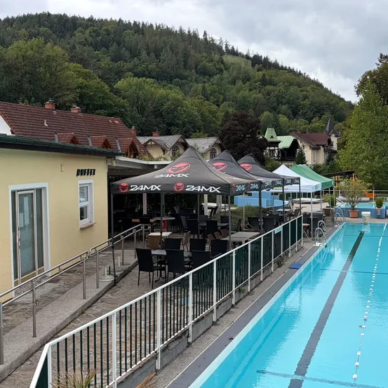Ein Freibadbereich mit einer Reihe schwarzer Zelte, Tischen und Stühlen, umgeben von einem Metallzaun. Ein Gebäude mit einem braunen Dach und Glastüren befindet sich in der Nähe.