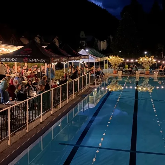 Eine nächtliche Szene eines Poolbereichs mit Menschen, die unter Zelten essen, umgeben von Bäumen und Lichtern.