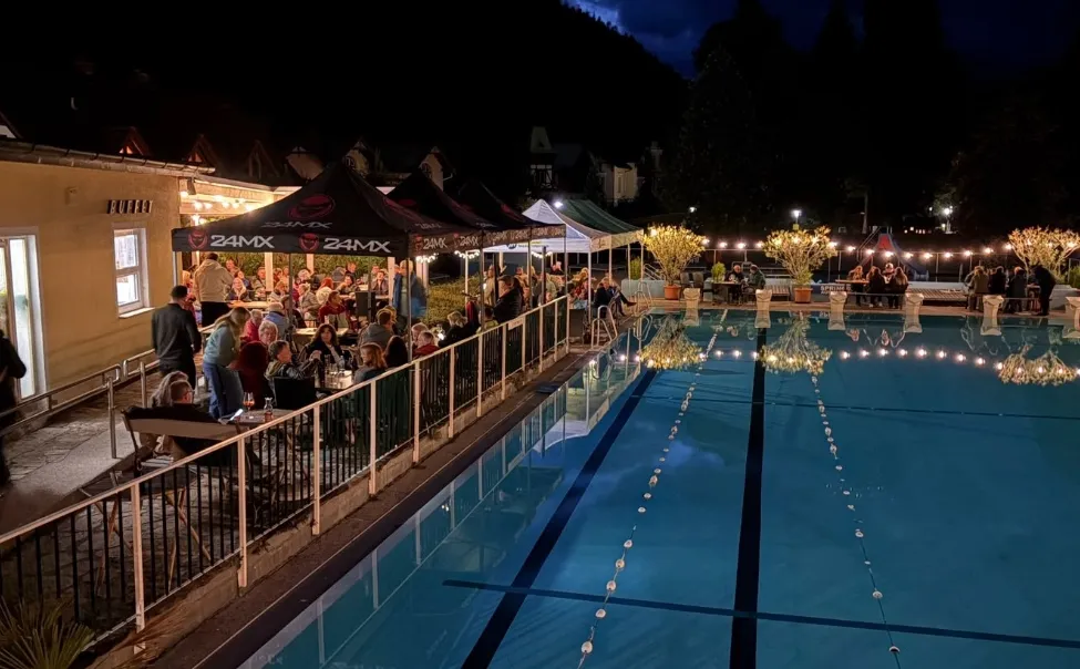 Eine nächtliche Szene eines Poolbereichs mit Menschen, die unter Zelten essen, umgeben von Bäumen und Lichtern.